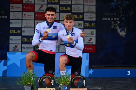 Zwei männliche Radfahrer in passenden weiß-blauen Trikots knien auf einem Podium und halten Medaillen in der Hand. Sie lächeln, vor ihnen stehen Trophäen und kleine Pflanzen. Im Hintergrund sind Sponsorenlogos zu sehen.