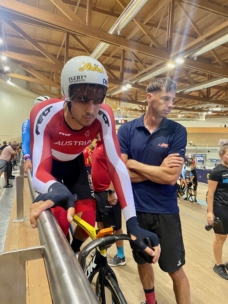 Ein österreichischer Radfahrer in einem rot-weißen Anzug und mit Helm bereitet sich auf ein Rennen in einem Indoor-Velodrom vor, wobei er den Fahrradlenker festhält, während ein Mann in einem marineblauen Hemd neben ihm steht. Im Hintergrund sind andere Menschen und Radfahrer zu sehen.