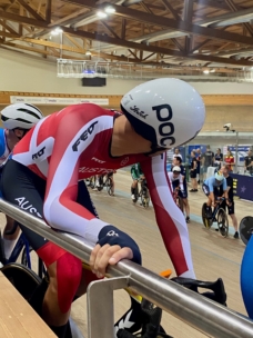 Ein Radfahrer in einem rot-weißen Rennanzug und einem aerodynamischen Helm hält sich an einem Metallgeländer in einem Indoor-Velodrom fest und bereitet sich auf ein Rennen vor. Im Hintergrund sind andere Radfahrer und Zuschauer zu sehen.