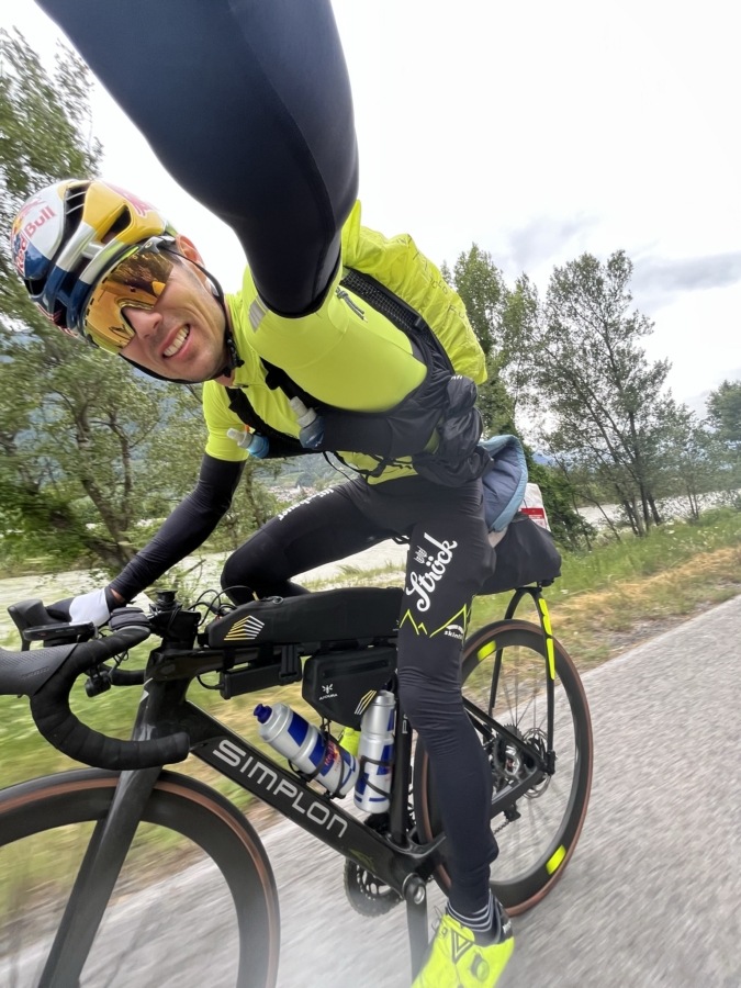 Ein Radfahrer mit gelbem Helm, Sonnenbrille und heller Kleidung macht ein Selfie, während er mit einem schwarzen Simplon-Fahrrad auf einer Landstraße fährt, mit Bäumen und bewölktem Himmel im Hintergrund.