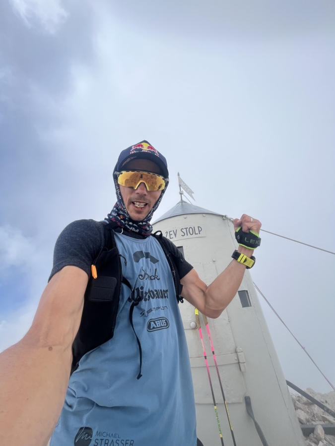 Ein Mann mit Sonnenbrille und Outdoor-Ausrüstung macht an einem nebligen Tag ein Selfie vor einer kleinen, weißen Berghütte mit der Aufschrift Triglavski Stolp. Er hält Trekkingstöcke in der Hand und lächelt in die Kamera.