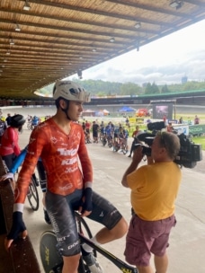 Ein Radfahrer in einem orange-roten Trikot und weißem Helm wird von einem Kameramann in einem gelben Hemd auf einem Outdoor-Velodrom gefilmt, während im Hintergrund andere Radfahrer und Veranstaltungszelte zu sehen sind.