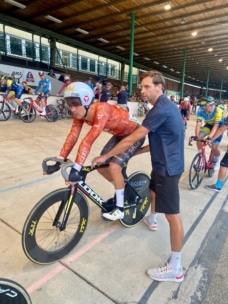 Ein Radfahrer in einem orangefarbenen Anzug und Helm wird von einem Trainer an der Startlinie eines Indoor-Velodroms festgehalten, während im Hintergrund andere Radfahrer warten.