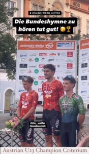 Drei männliche Radsportler stehen auf einem Podium mit Medaillen bei der österreichischen U23-Meisterschaft im Kriterium. Der mittlere Radfahrer steht auf der obersten Stufe, alle tragen passende Trikots. Auf einem Schild darüber steht: "Die Bundeshymne zu hören tut gut!".
