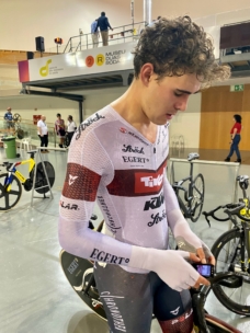 Ein junger männlicher Radfahrer im Rennanzug steht in einer Halle neben seinem Fahrrad und schaut auf ein Gerät in seinen Händen hinunter. Im Hintergrund der Radsportarena sind andere Fahrräder und Menschen zu sehen.