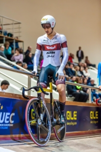 Ein Radfahrer in voller Rennmontur und mit Helm bereitet sich an der Startlinie eines Indoor-Velodroms vor und hält sich am Geländer fest. Die Zuschauer schauen von den Tribünen im Hintergrund zu.