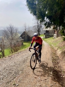 Ein Radfahrer in rotem Trikot und Helm fährt auf einem Kiesweg bergauf, der von Bäumen und rustikalen Häusern gesäumt ist, mit einer nebligen Landschaft im Hintergrund.