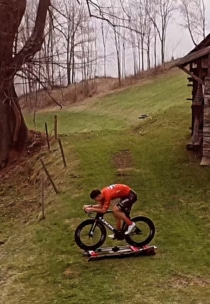 Ein Radfahrer in einem orangefarbenen Trikot fährt auf einem stationären Fahrradtrainer im Freien auf einem grasbewachsenen Hang in der Nähe eines rustikalen Holzgebäudes und blattloser Bäume.