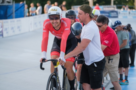 Ein Radfahrer in einem rot-schwarzen Trikot und Helm lächelt an der Startlinie, während ein Mann in einem weißen Hemd sein Fahrrad stützt. Mehrere andere Radfahrer und Offizielle stehen hinter ihnen auf einer Velodrombahn.