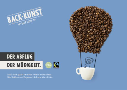 Ein Heißluftballon aus Kaffeebohnen erhebt sich aus einer weißen Kaffeetasse mit einer illustrierten Person darin. Der deutsche Text wirbt für fair gehandelten und biologisch angebauten Kaffee von Back Kunst und zeigt die Logos von Fairtrade und Bio. Blauer Hintergrund.