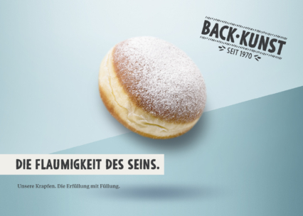 Ein rundes, mit Puderzucker bestreutes Gebäck (Krapfen) ist auf blauem Hintergrund mit deutschem Text abgebildet: Back-Kunst seit 1970 und Die Flaumigkeit des Seins. Unsere Krapfen. Die Erfüllung mit Füllung.