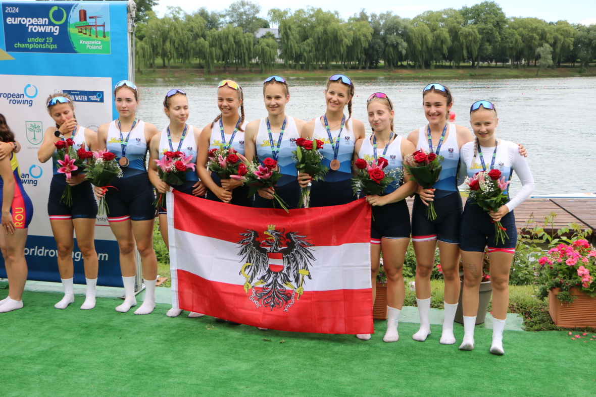 Acht Ruderinnen in passenden Uniformen stehen auf einer Wiese, halten Blumen und Medaillen in der Hand und tragen eine rot-weiß-rote Flagge mit einem schwarzen Adler. Im Hintergrund sind ein See und Bäume zu sehen, und links sind Veranstaltungsbanner zu sehen.