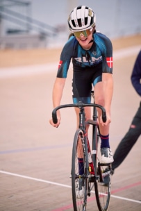 Ein Radfahrer, der einen Helm, eine Sonnenbrille und einen blauen Rennanzug mit dänischen Flaggen an den Ärmeln trägt, fährt auf einem Bahnrad in einer Radrennhalle.