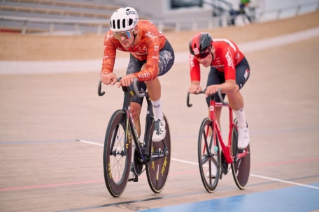 Zwei Radfahrer in Rennkleidung und mit Helm treten auf einer Radrennbahn gegeneinander an. Der Radfahrer im Vordergrund trägt ein orangefarbenes Trikot, der hintere ein rotes. Beide fahren auf aerodynamischen Fahrrädern mit hoher Geschwindigkeit.