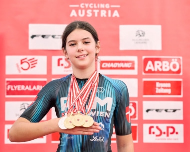 Eine junge Radfahrerin im blau-schwarzen Trikot hält vier Goldmedaillen in die Höhe und lächelt vor einem roten Hintergrund mit dem Schriftzug "Cycling Austria" und mehreren Sponsorenlogos.