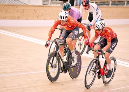 Vier Radfahrer fahren ein Rennen auf einer Indoor-Velodrombahn. Zwei Radfahrer in orangefarbenen Trikots führen dicht beieinander, während zwei Konkurrenten in weißen und blauen Trikots hinterherfahren. Die Holzbahn und die Helme sind sichtbar.