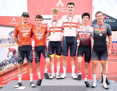 Sechs männliche Radfahrer mit Medaillen stehen auf einem Podium und lächeln in die Kamera. Hinter ihnen befindet sich ein roter Hintergrund mit Logos von Cycling Austria und Sponsoren. Einige tragen passende Trikots, andere tragen unterschiedliche Team-Outfits.