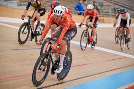 Radfahrer mit Helmen und orangefarbenen Trikots fahren ein Rennen auf einer hölzernen Indoor-Velodrombahn, wobei einige Fahrer im Hintergrund dicht hinter ihnen herfahren. Die Plätze in der Arena sind größtenteils leer.