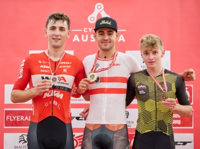 Drei männliche Radsportler stehen auf einem Podium, jeder trägt eine Medaille um den Hals. Sie lächeln, halten sich in den Armen und stehen vor einem roten Hintergrund mit Veranstaltungslogos.
