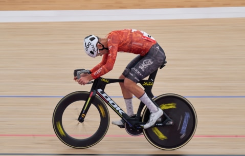 Ein Radfahrer in einem orange-schwarzen Rennanzug und weißem Helm fährt auf einem aerodynamischen Bahnrad mit hoher Geschwindigkeit auf einem Indoor-Velodrom, wobei der Hintergrund unscharf ist, um die Bewegung zu zeigen.