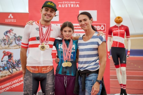 Drei Personen stehen lächelnd bei einer Radsportveranstaltung: ein Mann und ein Mädchen im Teenageralter, beide tragen Medaillen, und eine Frau. Im Hintergrund ist eine Schaufensterpuppe in Radfahrerkleidung mit einem Kürbiskopf und Veranstaltungsbannern zu sehen.