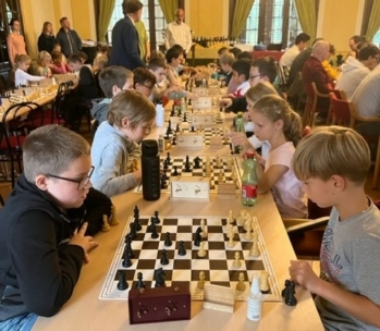 Kinder und Erwachsene sitzen sich an langen Tischen gegenüber und spielen in einem hellen, überfüllten Raum konzentriert Schach. Schachbretter, Uhren und Getränke stehen auf den Tischen. Im Hintergrund stehen Zuschauer.