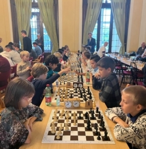 An mehreren Tischen sitzen sich Kinder gegenüber, die in einem hellen Raum mit großen Fenstern und grünen Vorhängen konzentriert Schach spielen. Schachuhren und Limonadenflaschen sind auf den Tischen zu sehen.
