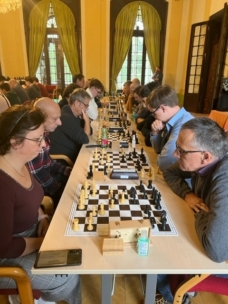 An einem langen Tisch in einem gut beleuchteten Raum mit großen Fenstern, grünen Vorhängen und klassischem Dekor sitzen sich mehrere Paare gegenüber und beschäftigen sich intensiv mit Schachspielen.