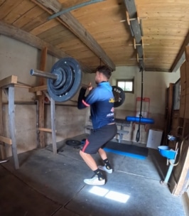 Eine Person in Sportkleidung und Turnschuhen hebt eine Langhantel mit Gewichten in einer rustikalen Holzhalle mit freiliegenden Balken und einfachen Trainingsgeräten.