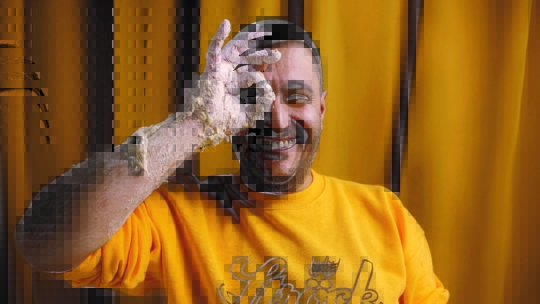 Ein lächelnder Mann in einem gelben Ströck-Sweatshirt hält eine mit Knete bedeckte Hand hoch und macht ein "OK"-Zeichen über sein Auge, während er vor einem passenden gelben Vorhang steht.
