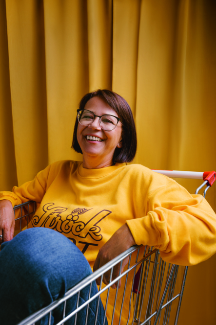 Eine Frau mit Brille, die ein gelbes Sweatshirt und blaue Jeans trägt, lächelt, während sie entspannt in einem Einkaufswagen vor dem Hintergrund eines gelben Vorhangs sitzt.