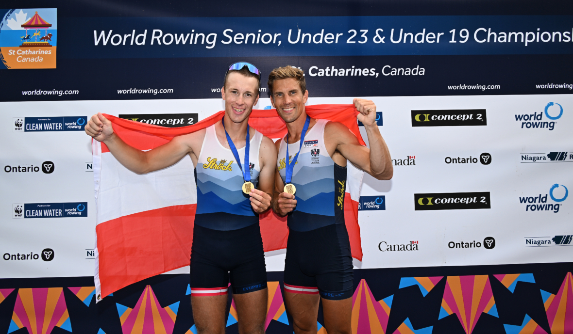 Zwei männliche Ruderer in passenden Uniformen halten Goldmedaillen und eine rot-weiße Flagge und lächeln vor einer Kulisse mit der Aufschrift World Rowing Senior, Under 23 & Under 19 Championships, St. Catharines, Kanada.