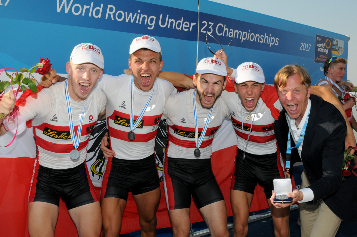 Fünf männliche Athleten in Ruderuniformen und mit Medaillen feiern mit einem Trainer vor einem "World Rowing Under 23 Championships"-Banner, lächelnd und mit Blumen in der Hand, mit einer rot-weißen Flagge im Hintergrund.