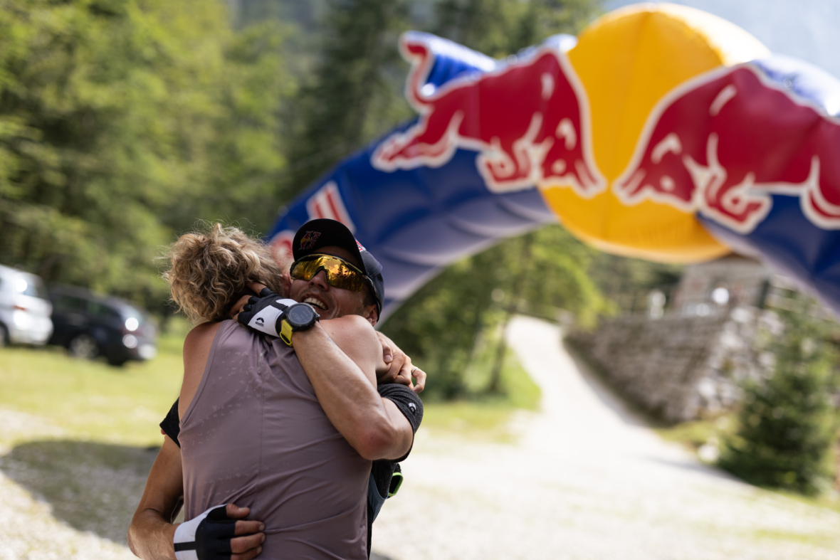 Zwei Athleten umarmen sich feierlich in der Nähe eines aufblasbaren Bogens der Marke Red Bull im Freien, während im Hintergrund an einem sonnigen Tag Bäume und ein Kiesweg zu sehen sind.