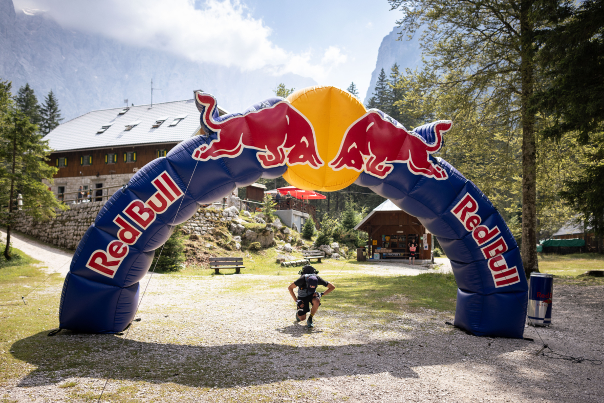 Eine Person mit einem Rucksack hockt unter einem großen aufblasbaren Red Bull-Bogen in einem bergigen Außenbereich mit Bäumen und Gebäuden im Hintergrund.