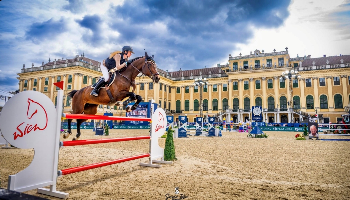 Ein Reiter und sein Pferd springen während eines Reitturniers über einen rot-weißen Sprung, mit einem großen historischen Gebäude und einem dramatischen Wolkenhimmel im Hintergrund. Zuschauer und Veranstaltungsschilder sind zu sehen.