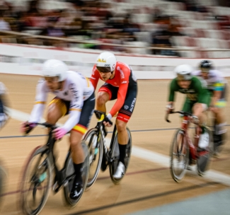 Vier Radfahrer in Rennkleidung und Helmen treten auf einer Indoor-Radrennbahn an, die in Bewegungsunschärfe aufgenommen wurde. Der Fahrer im roten Trikot befindet sich in der Mitte und folgt dem führenden Fahrer.