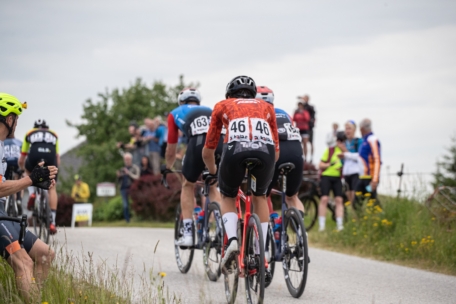 Die Radfahrer, die Helme und nummerierte Trikots tragen, fahren auf einer gepflasterten Straße bergauf, umgeben von Gras und Wildblumen, während Zuschauer vom Straßenrand aus bei bewölktem Himmel zusehen.