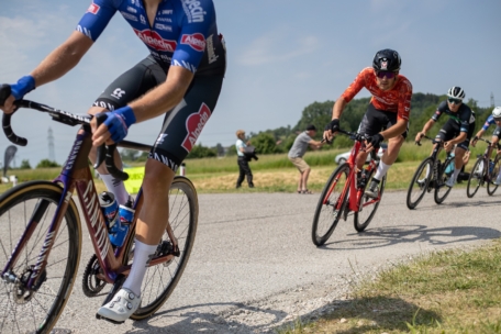 Eine Gruppe von Radfahrern in farbenfroher Rennkleidung fährt auf einer sonnigen Straße im Freien um eine Kurve, während im Hintergrund Zuschauer und Grünflächen zu sehen sind.