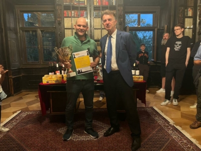 Zwei Männer stehen in einem Raum; einer hält eine große Trophäe und eine Urkunde in der Hand. Hinter ihnen sind ein Tisch mit Auszeichnungen, Bücherregale und mehrere Personen zu sehen. Die Umgebung wirkt formell und feierlich.