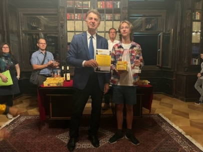 Ein Mann im Anzug und ein Jugendlicher in Freizeitkleidung stehen Seite an Seite mit Urkunden und Trophäen in einer holzgetäfelten Bibliothek, während im Hintergrund mehrere Personen zusehen.