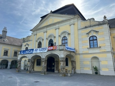 Ein prächtiges gelbes Gebäude mit verzierten Säulen und Statuen am Eingang, gewölbten Fenstern und Fahnen, die über dem Eingang hängen, unter einem sonnigen blauen Himmel.