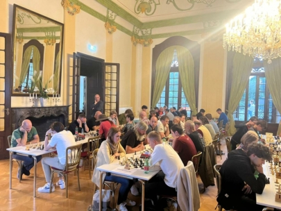 In einem geräumigen, eleganten Raum mit hohen Decken, Kronleuchtern und großen, mit grünen Vorhängen verhangenen Fenstern wird konzentriert Schach gespielt. Mehrere Tische sind mit Schachbrettern aufgestellt und die Spieler konzentrieren sich auf ihr Spiel.