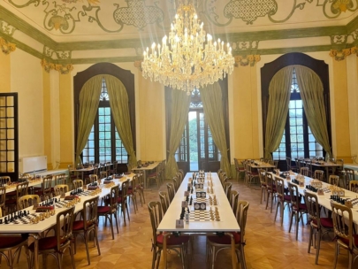 Eleganter Saal mit hohen Decken, einem großen Kronleuchter und hohen Fenstern mit grünen Vorhängen. Mehrere Tische sind für ein Schachturnier aufgestellt, jeder mit Schachbrettern und Stühlen, die ordentlich angeordnet sind.