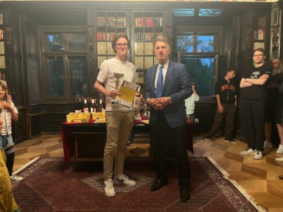 Ein junger Mann mit einer Trophäe und einer Urkunde steht neben einem älteren Mann im Anzug. Sie befinden sich in einer reich verzierten Bibliothek mit Bücherregalen und einem roten Tisch mit Preisen. Mehrere Personen stehen im Hintergrund und beobachten.