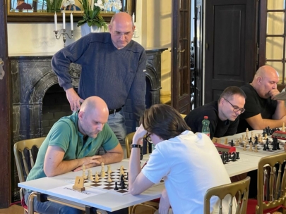 Mehrere Männer sitzen an Tischen mit Schachbrettern und konzentrieren sich auf eine Schachpartie bei einem Turnier. Ein Mann steht und beobachtet die Partien, während andere auf ihre Züge konzentriert sind. Die Umgebung ist reich verziert, mit elegantem Dekor und einem Kamin.