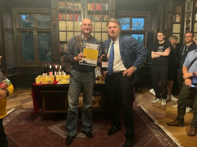 Zwei Männer stehen in der Mitte eines Raumes; einer hält eine Trophäe und eine Urkunde in der Hand, während der andere in ein Mikrofon spricht. Um sie herum stehen Menschen, und im Hintergrund steht ein Tisch mit Weinflaschen und Kisten. Bücherregale säumen die Wände.