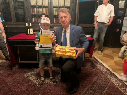 Ein kleiner Junge mit einer Urkunde und einem Pokal steht neben einem älteren Mann in einem blauen Anzug, der kniet und eine Schachtel mit der Aufschrift Schoko-Bananen in der Hand hält. Hinter ihnen sind Menschen in einem Raum mit Bücherregalen und einem roten Teppich versammelt.