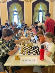 Kinder und Jugendliche spielen Schach an langen Tischen in einem großen, gut beleuchteten Raum mit hohen Decken und großen Fenstern. Zuschauer und Eltern stehen herum und beobachten die Spiele. Ein Schild mit der Aufschrift JUGENDLICHE steht am nächsten Tisch.