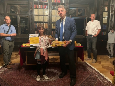 Ein junges Mädchen steht neben einem Mann im Anzug, beide halten Auszeichnungen, in einem Bibliotheksraum mit Holzregalen und Büchern. Drei weitere Erwachsene und ein Kind stehen im Hintergrund und beobachten die Szene.